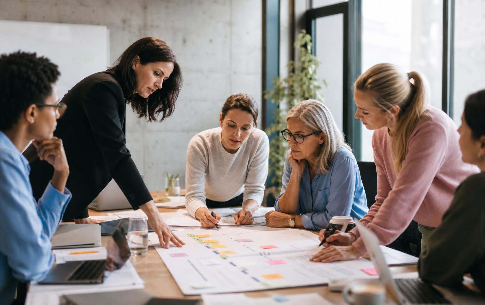 A diverse team of professionals, mostly women, working together on a project recovery plan around a table with documents and sticky notes